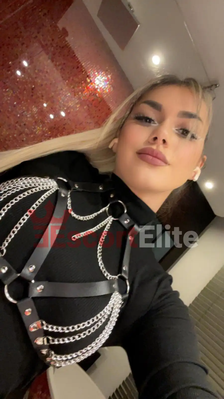 Blonde Escort Wien Jasmin mit schwarzem Leder-Harness und Silberketten, glamouröses Porträt